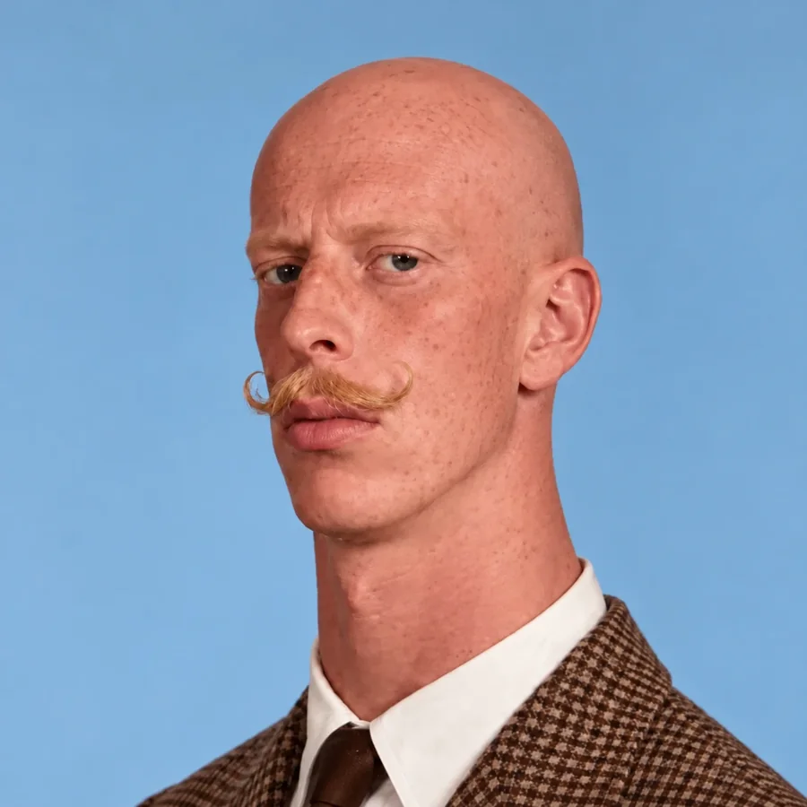 A bald man with a curled ginger mustache and a tweed jacket, looking unimpressed